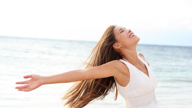 Happy woman in white dress smiling with open arms by the ocean, symbolizing freedom, confidence, and positive transformation through hypnosis