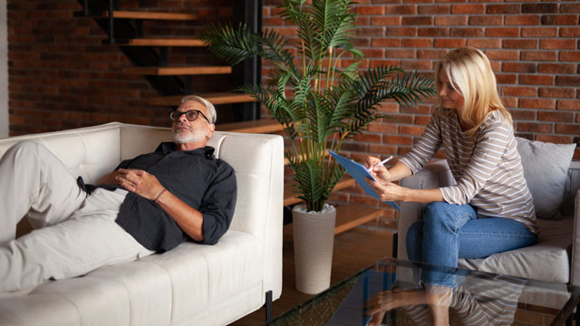 Hypnosis session with mature man relaxing on sofa while female therapist takes notes in a calm office